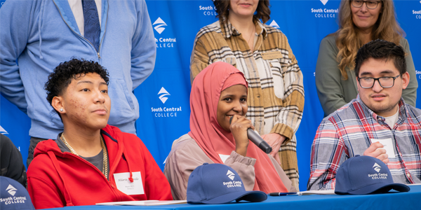 Signing Day students on the Faribault campus grouped together smiling and wearing SCC caps