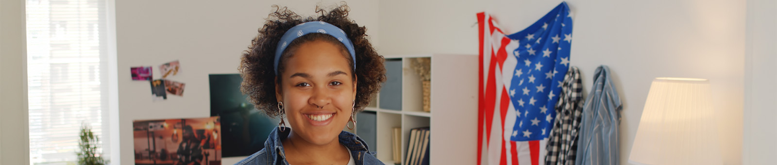 College student standing in apartment with American Flag draped across wall smiling at camera