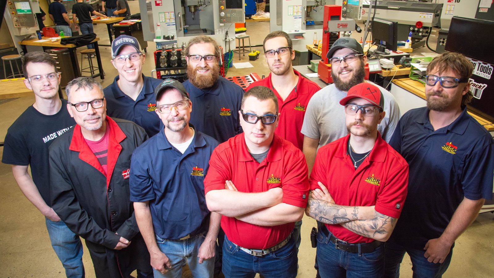 A team of machine tool technicians in company shirts and safety glasses gather in a college machining lab and look up at the camera.