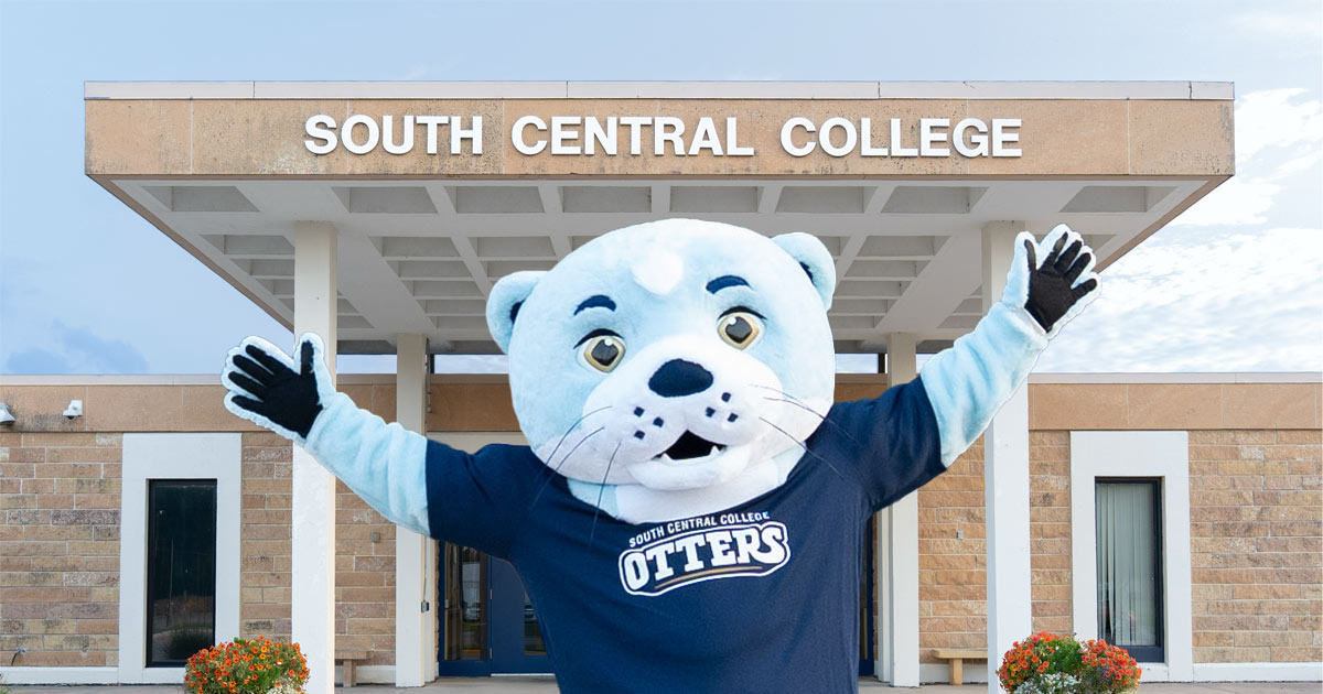 A person in a Splash costume, SCC's otter mascot, stretching out it's arms in front of the entryway to South Central College.